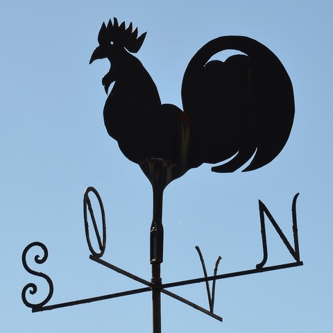 A photo of a weather vane against a clear blue sky. There is a flat metal rooster on top, and the directions are marked with the letters N, Ø, S, and V.