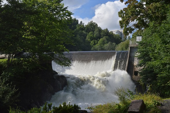 A photo of water flowing over a dam. It is surrounded by trees. The sky is blue with white clouds in it.