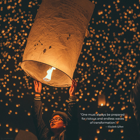 A woman in the process of letting go of a large, lit lantern, letting it float into the black sky with countless other lanterns seen in the background. The quote on the photo reads, â€œOne must always be prepared for riotous and endless waves of transformation.â€� by Elizabeth Gilbert.