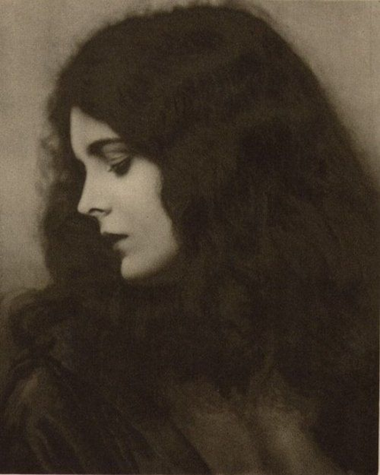 Black and white photo of a woman in profile. She has long, dark hair and turns her head away from the camera, looking down. 