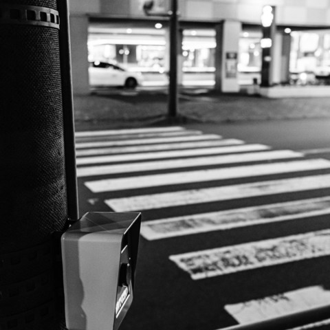A black and white photo taken from a pedestrian's perspective at a crosswalk, with the focus directed towards the button on the pole. The image captures the intersection of pedestrian sight and the mechanism that governs their movement.