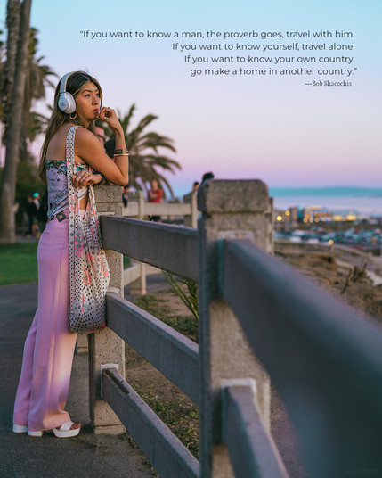 A woman stands at a fenced viewpoint, overlooking a city's coastal area. The quote on the photo reads, "If you want to know a man, the proverb goes, travel with him. If you want to know yourself, travel alone. If you want to know your own country, go make a home in another country." by Bob Shacochis.