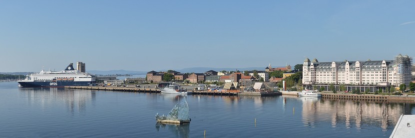 A panoramic photo of a bay with a large building to the right and a large ship to the left of the far side of the bay. There is a sculpture in the middle of the bay. The sky is blue and cloudless.