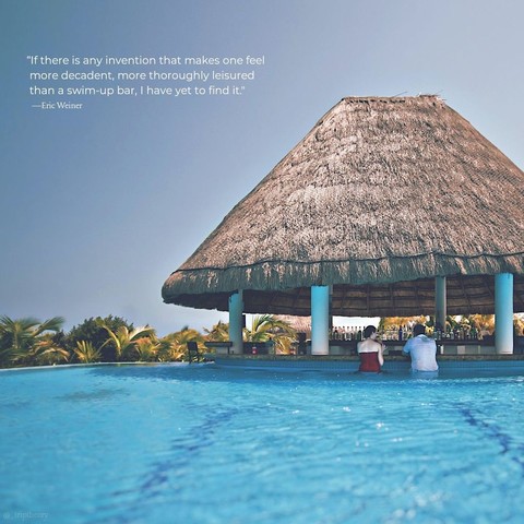 Two people sit on underwater stools at a swim up bar, covered by a large thatched roof. The quote on the photo reads, "If there is any invention that makes one feel more decadent, more thoroughly leisured than a swim-up bar, I have yet to find it." by Eric Weiner.