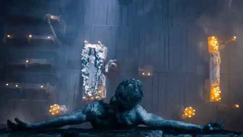 GIF from the 1967 film 'Viy'. It shows the camera looking down from above a dark statue of Christ on the cross. Below, a vigil is set up, with a beautiful pale, dark haired girl in white laid in a coffin, which is surrounded by candles and flowers. By her side, an older man looks over her corpse. 