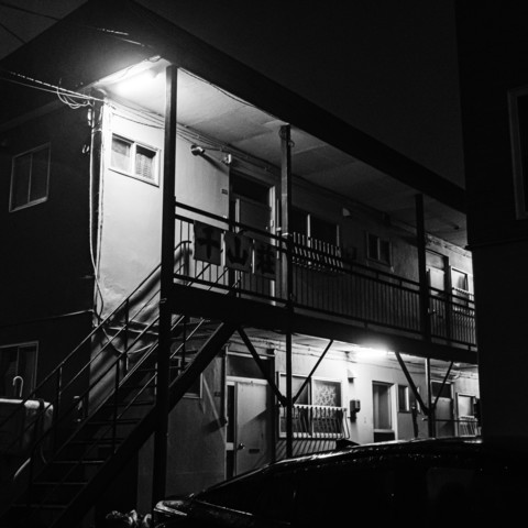 A black and white photograph of a two-story apartment in the dead of night. The sparse outdoor lighting makes it difficult to discern even the keyhole on the door.