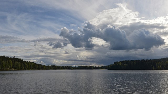 A photo of a lake bordered by conifers. The sky is filled with clouds.