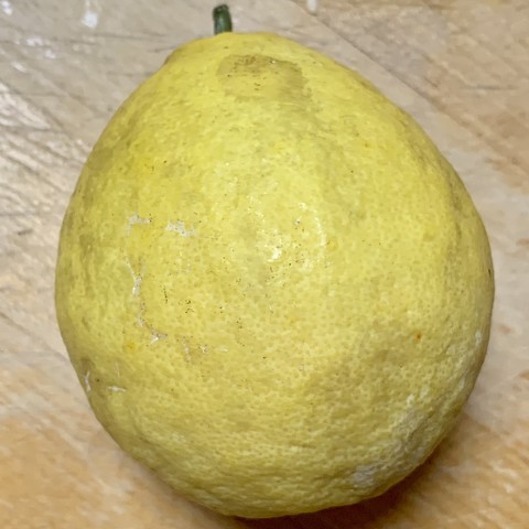 A round yellow fruit that is slightly pointed at the green stem. It is laying on a wooden countertop. It is smaller and rounder than a standard grapefruit. 