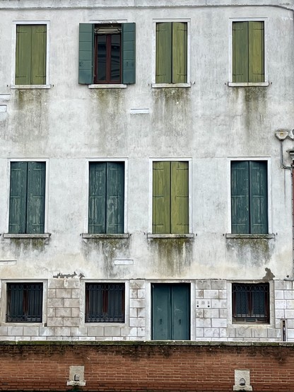 Colour photo of a dingy white three storey building, with a terracotta brick wall in front. On the ground floor are three regularly spaced identical square windows, dark almost black with iron bars, with a closed teal coloured door breaking up the symmetry second from the right. First floor has long rectangular windows, three with closed shutters painted teal, apart from the one above the door which is a mossy pale green. The top floor has smaller rectangular shuttered windows, three painted the mossy colour, apart from the second from the left which has its shutters open. There are streaks below each of the window ledges, like the fading strokes from a paintbrush. The overall effect of the image is of an abstract cubist painting. 
