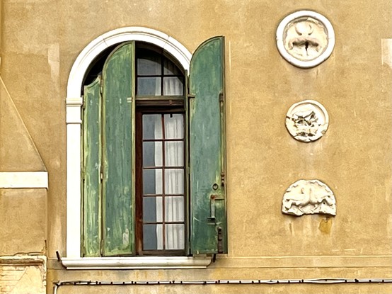 Colour photo of a sandy-ochre coloured wall, with, on the right of the image three white, worn, circular medallion ornaments, and on the left, a white-framed arched window with peeling moss-green shutters, one of which is open and the other closed, giving us a view of half a pane of mullioned glass with a white curtain behind. To balance out the composition, there is a white strip of plaster a third of the way up the image on the far left side, and a white line of cable running across the bottom of the picture. 