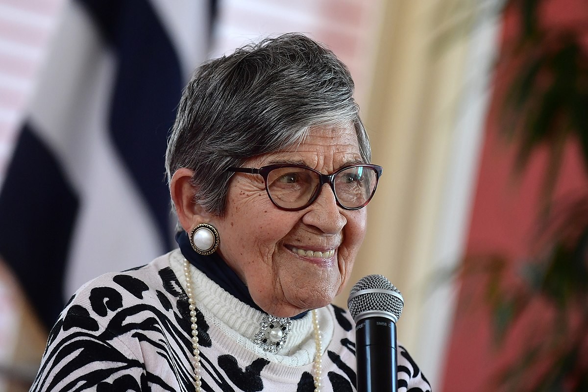 A French Jewish woman, Ginette Kolinka. An older lady in glasses, large earrings, black & white patterned sweater. She is smiling and holding a microphone.