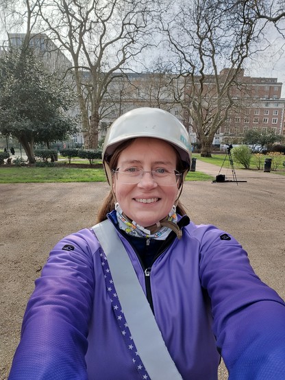 A middle aged white woman in a pale gold cycle helmet, purple jacket and reflective sash 