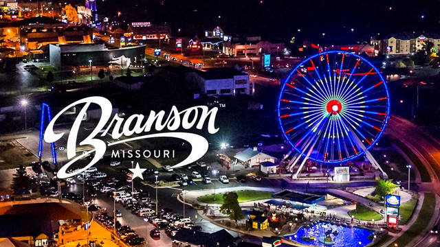 A postcard-like image of Branson, Missouri, at night from above, featuring a monumental, lit-up, white -- all-white, come to think of it -- "Branson, Missouri" sign and a Ferris wheel lit up in red, white, and blue beside it. Kind of pretty, actually.