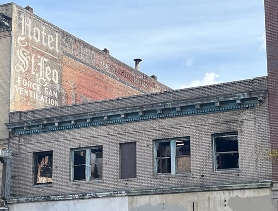 The second floor of a vacant old building with empty, broken windows. Faintly painted on the wall of an old brick building beside it and a story taller is HOTEL ST. LEO - FORCE FAN VENTILATION.