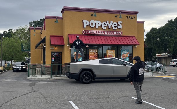 Fanboy with a cellphone photographing a second man with his arms outstretched standing on top of a Cybertruck in front of a Popeye’s Louisiana Kitchen 