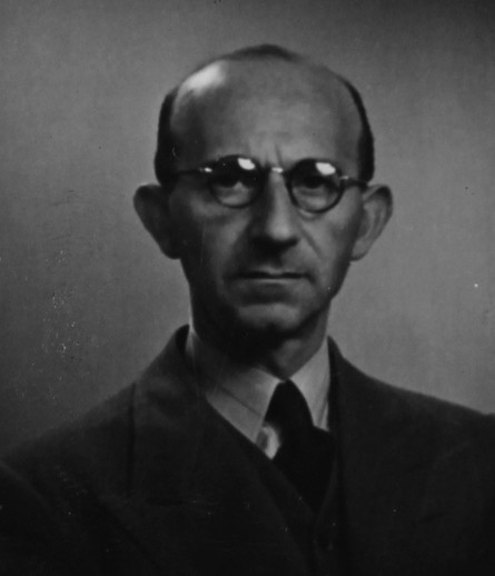 A photo of the face of a mature man in round-framed glasses. He is wearing a suit. He has bold sides of his head. 