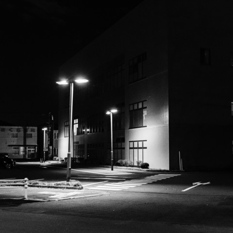 This is a black and white image capturing a night scene of a quiet and empty street. Two tall streetlights stand out, casting bright light over the area. One side of the image shows a building with several unlit windows. The ground features a pedestrian crossing marked with white lines. There are no visible people or moving vehicles, creating an empty, tranquil scene. The shadows from the lights produce a contrast between the illuminated and dark areas.