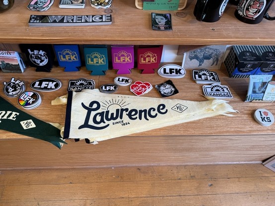 Various knick-knacks at the book store that are Lawrence KS themed. Stickers, flags, and cup holders with “Lawrence KS” printed on them can be seen. 