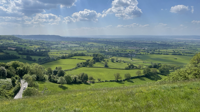 View from Coaley Peak across the Severn Vale.  Field of green out towards the river with Frocester Hill road in the foreground 