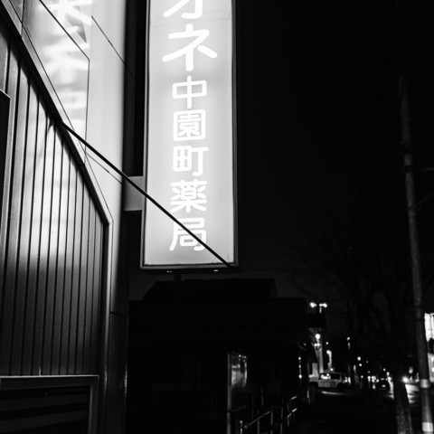 This is a black and white photograph taken at night. The image captures a portion of a building facade and a vertically oriented illuminated sign. The sign is glowing brightly, contrasting against the dark night sky and the dimly lit street in the background. The building's exterior features vertical panels, adding texture to the scene. A narrow street is visible on the right side of the photograph, with a few indistinct lights and silhouettes of trees and structures in the distance. The overall atmosphere is quiet and slightly eerie, with sharp contrasts between light and shadow.