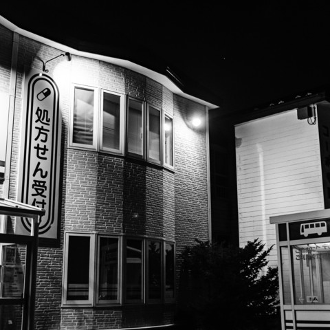 The black and white photograph captures a nighttime scene of a building. The building has a modern architectural style with a facade designed to resemble bricks, though it is not made of actual bricks. The photograph is taken from an upward angle, showcasing the side of the building which includes several windows. There is a prominent, vertically oriented sign attached to the building. Additionally, a bright light illuminates the wall above the sign. To the right of the building, there is a bus stop structure with a transparent shelter, partially visible.