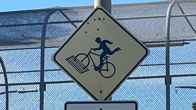 A yellow road sign with a silhouette of a bicyclist's front wheel falling into a grate, with the rider about to go flying