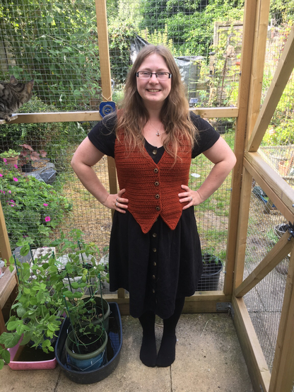40 year old woman wear a rust coloured crochet waistcoat over a black buttoned dress. She is smiling and has her hands on her hips. She has long straggly blonde/light brown hair. She is standing in a catio with an overgrown garden in the background.