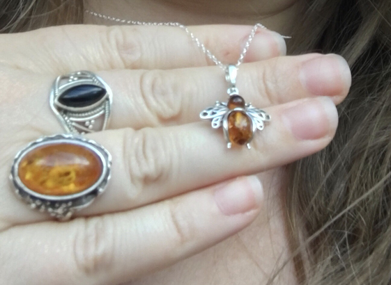Silver and amber bee necklace, showcased on hand that wears two silver rings, one has a large amber stone and the other a back stone.