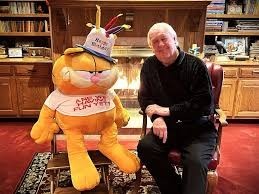 Cartoonist Jim Davis sitting next to an oversized Garfield plush wearing a T-shirt that reads "Are we having fun yet?"