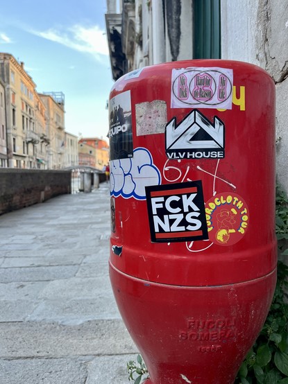 Colour photo of a bright red fire hydrant covered in stickers, the most prominent of which says FCK NZS in white letters with red lines above and below on a black background. Behind the hydrant a typically Venetian street, with stone flagging, a canal-side wall, and houses coloured blush-pink in the sun, runs into the distance. 
