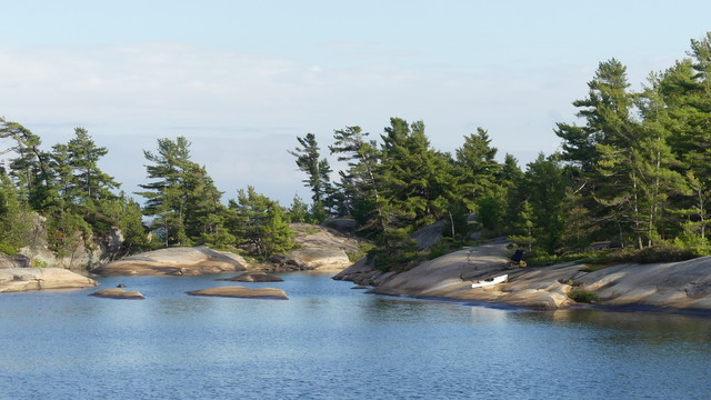 A white kayak sits on smooth brown rock topped with pines make the shoreline of a rippled cove under blue sky.  More rocks emerge from the cove.