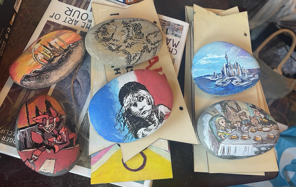 Six pebbles, one painted to show the landscape of Gallifrey, one a map of Middle Earth, the city of Atlantis from Stargate Atlantis, Alastair from Hazbin Hotel, the Cosette logo from Les Misérables, and a copy of Tenniel’s illustration of the Mad Hatter’s tea party. Each stone is about three inches by two.