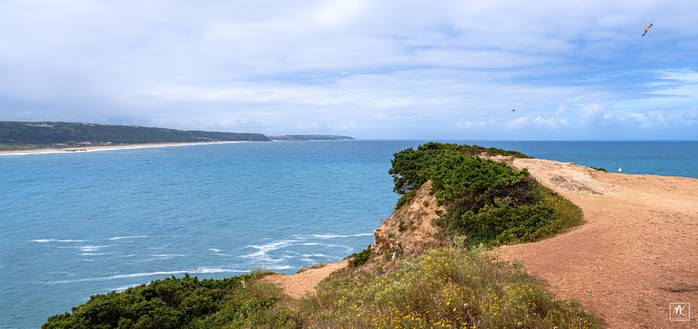 Color photo taken from the top of a coastal cliff looking out across a bay towards another shore with a mostly cloudy sky above. 