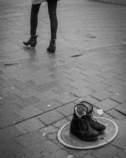 Sidewalk. Woman walking away to upper left. Only her legs and booted feet showing. Foreground is a pair of abandoned winter boots on an electrical access panel. Black and white.