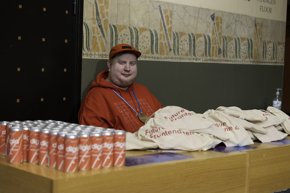 Juhis sitting behind a desk wearing bright red cap and hoodie and a conference badge around his neck. On the desk, a bunch of energy drink cans and tote bags.