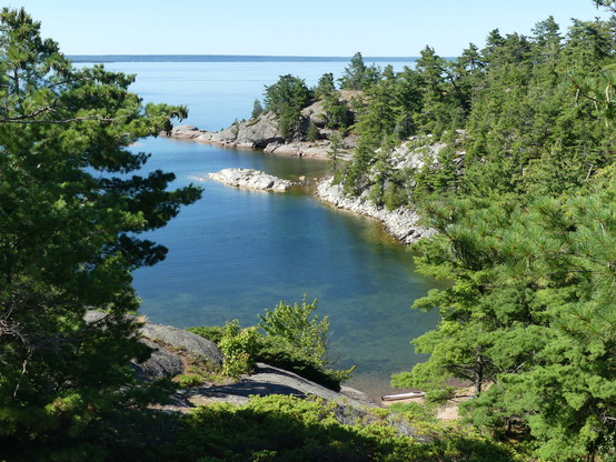 A cove with a shoreline of smooth bedrock and pine trees on calm water on a cloudless day.  The picture is taken from a height and rocks are visible on the bottom of the cove.  A white kayak is beached on sand at the near end of the cove.