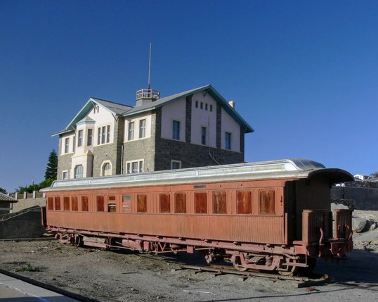 A dusty ocher red railroad car stands before a big stone and white building with lots of different sized windows. The sky is blue and empty. The scene feels rather desolate. This is the train station of Lüderitz, Namibia in 2011.