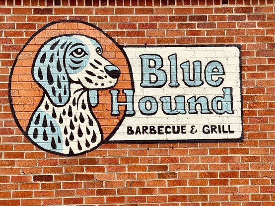 A mural painted on a brick wall features an illustration of a blue and white hound dog with black spots, set against an orange circle background. Next to the dog, the text reads 