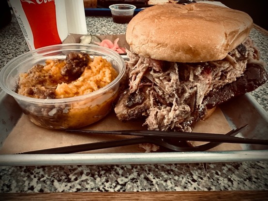 A tray that includes a large sandwich called the SQUATCH-WICH with pork, brisket, turkey, and slaw on a toasted bun. Next to the sandwich is a plastic container of mashed sweet potato crunch. There is a fork and knife on the tray, and a drink cup in the background. The setting is a the Blue Hound Barbecue & Grill in Dillard, GA.