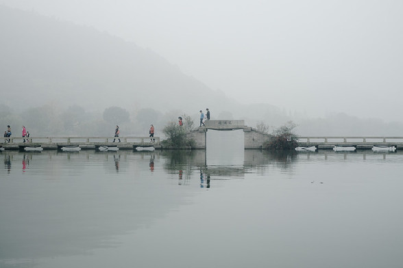 Eight or nine people, just small figures in the distance, walk across a low stone bridge spanning a mist-covered river, with misty hills and trees just vaguely visible in the background. 