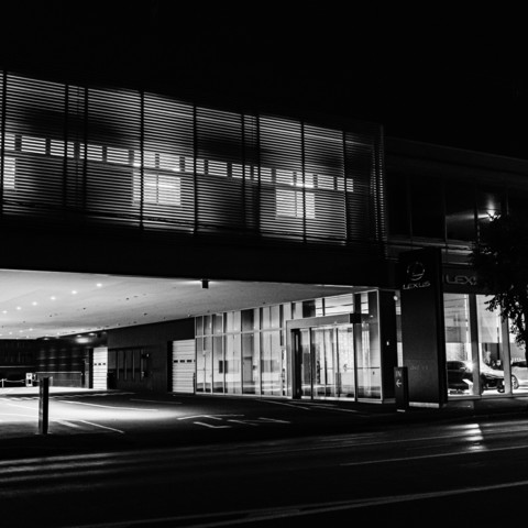 This black and white photograph shows an automobile dealership at night after closing. The building features a modern architectural design with extensive use of glass and metal. The main entrance area is well-lit, with overhead lights illuminating the exterior. Large windows display the interior, showing a showroom with vehicles visible inside. The dealership has a prominent awning extending over a driveway, creating a covered entrance. The signage indicates the brand, though it's not readable in this description. The surroundings are dark, with minimal light sources other than the dealership's own lighting.