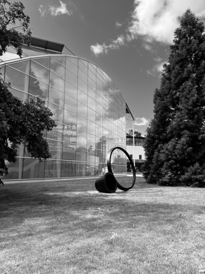 A black and white photo of The Squire Library at the University of Cambridge, the foreground of the photo is a green lawn then the building is construed glass windows which are reflecting the sky with a few fluffy clouds. There is a large evergreen tree on the right side of the photo and a metal sculpture in front of it and in the middle of the frame.