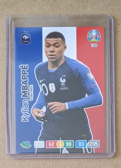 A trading card in a toploader shows Mbappé with a dark blue jersey and long blue sleeves. A French flag is used as a background