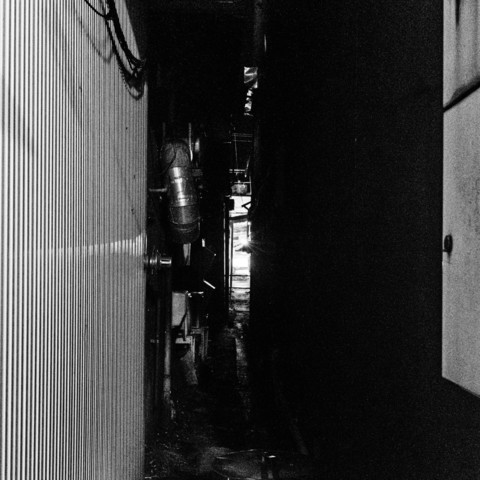 The photograph depicts a narrow alleyway between two buildings. The image is in black and white, with high contrast and heavy shadows. On the left side, the wall features vertical corrugated metal panels, which create a distinct linear pattern. The wall also has some pipes and ductwork attached to it, with one prominent cylindrical duct running horizontally.In the center of the image, there is a narrow pathway leading to a bright light source at the far end, which contrasts starkly with the surrounding darkness. The right side of the alleyway is mostly in shadow, making it difficult to distinguish any details. The overall atmosphere of the photograph is dark and somewhat ominous, with the light at the end providing a focal point that draws the viewer's attention.