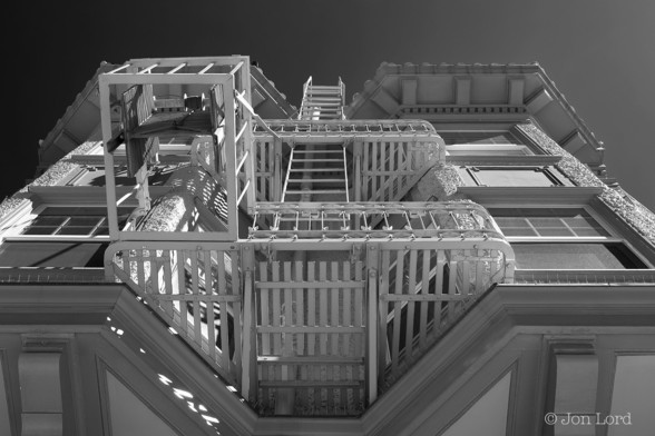 An Abstract Black And White Photo In Landscape Format. In The Image The Camera Is Pointed Vertically Upwards Parallel To The Front Of A Period San Franciscan House. In The Centre Of The Photo Are The Metal Balconies And Fire Escapes. On Either Side Are Bay Windows. Above The Two Floors That Are In View Are Large Eves That Overhand The Facade By About A Metre. Beyond The Roof Is A Clear And Cloudless Sky, Darkened To Near Black Using A Red Filter.
There Is No One Visible In The Photo.

Nob Hill, San Francisco. 2016