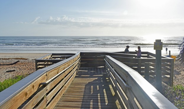 Beach dune boardwalk with zig zag accessible ramp to sandy beach with blue water and cloudy sky with sun glare on right and silhouettes of man and woman on ramp