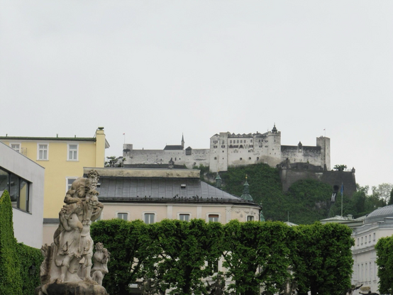Hohensalzburg fortress sits high above the city of Salzburg. Photo taken from Mirabell Garden. One of the statue from the garden is at the forefront to the left of the photo. A row of tall green bushes stand behind the statue. A round dark roof is seen between the bushes and the fort.  