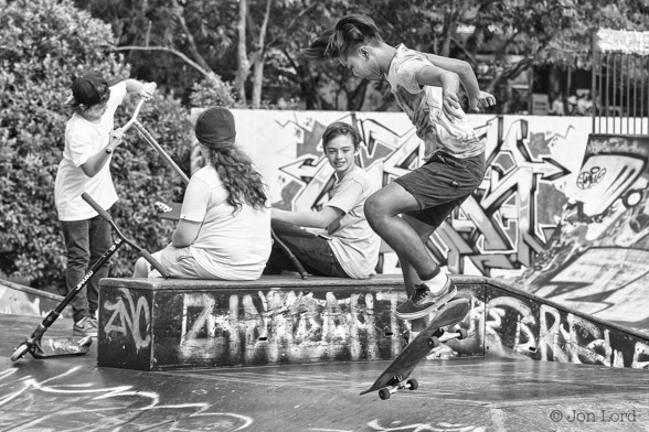 A Black And White Photo In Landscape Format. The Image Is Focused On A Teen: Airborne, His Skateboard - Also Airborne, Just A Few cm Below His Feet. Behind Are Is A Small Group Of Teens With One Watching. Surrounding The Group Is Graffiti Sprayed On The Walls. There Is Vegetation Beyond The Walls. The Locations Is An Urban Skateboard Park In Singapore. 2015