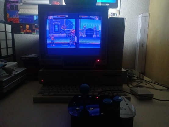Un Amiga 1000 et son écran affichent un beat'n all dans une ambiance cosy. Un joystick type arcade est posé devant l'ordinateur. 