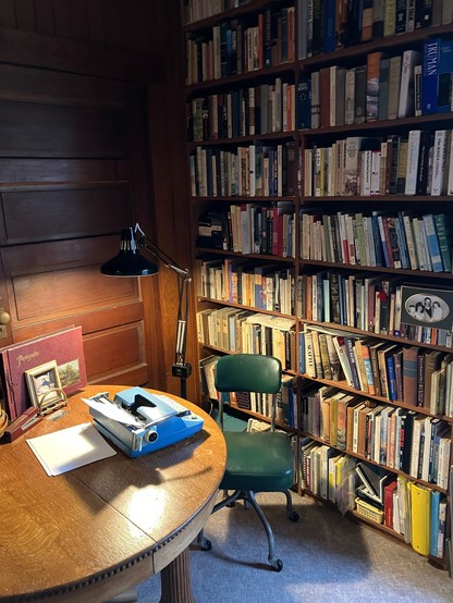A small table in a room with a wall of wood and a wall of books. There is an old typewriter on the table and an old office chair beside it. 